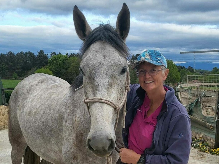 Emily smiles at the camera, holding the reigns to a grey horse in an outdoor setting.