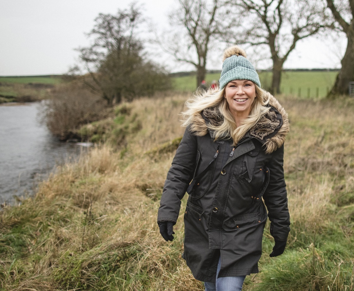 A woman in her thirties to forties and wearing a green beanie hat, smiles at the camera walking outdoors in the countryside.