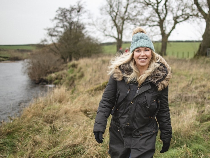 A woman in her thirties to forties and wearing a green beanie hat, smiles at the camera walking outdoors in the countryside.