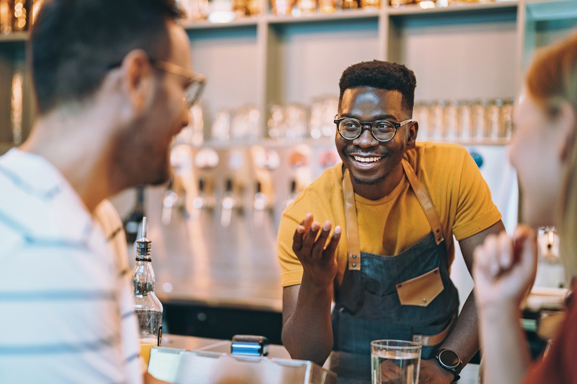 A barman talks to customers over a brightly lit bar.