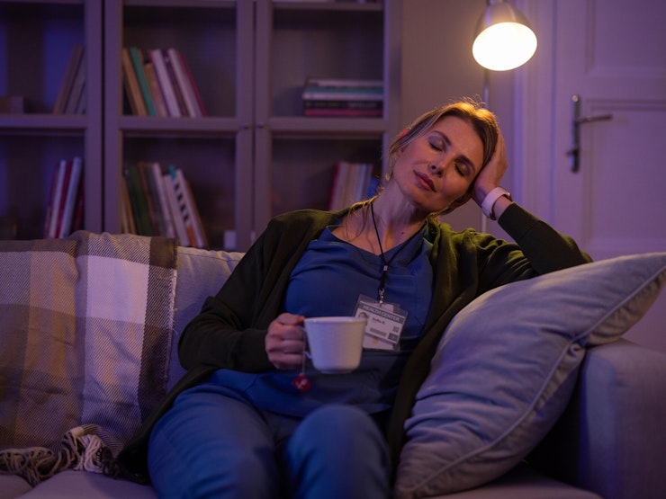 A woman in scrubs holds a cup of tea and supports her head and closes her eyes while sitting on a sofa in a darkened room.