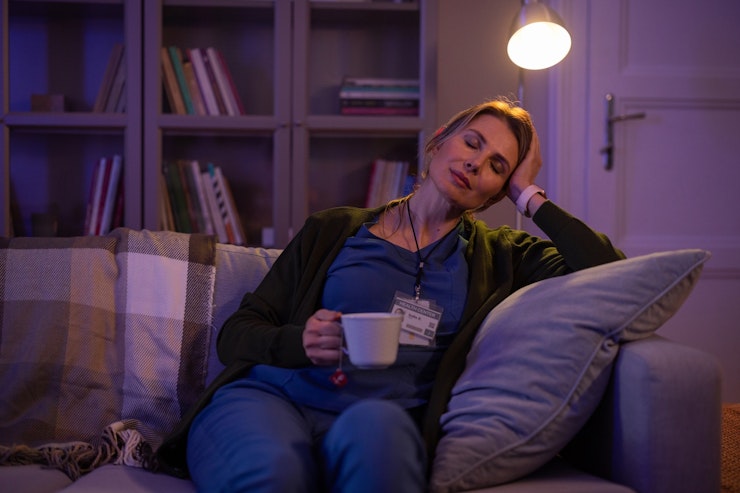 A woman in scrubs holds a cup of tea and supports her head and closes her eyes while sitting on a sofa in a darkened room.