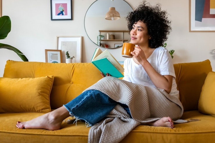 A woman with a black afro reads a book while drinking tea from an orange mug as she lounges on the sofa.