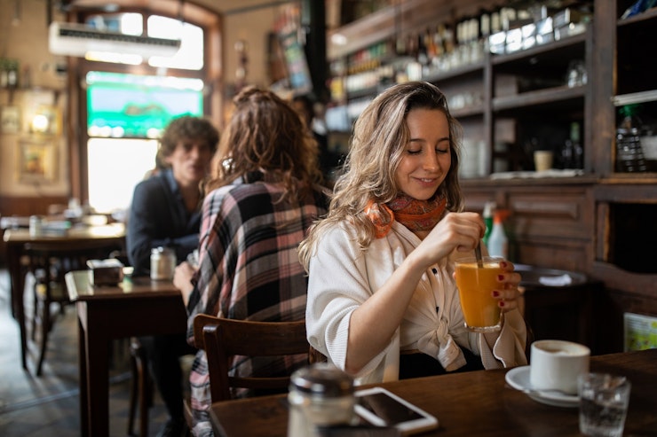 A woman with blonde wavy hair sitting in a café smiles as she stirs an orange drink.