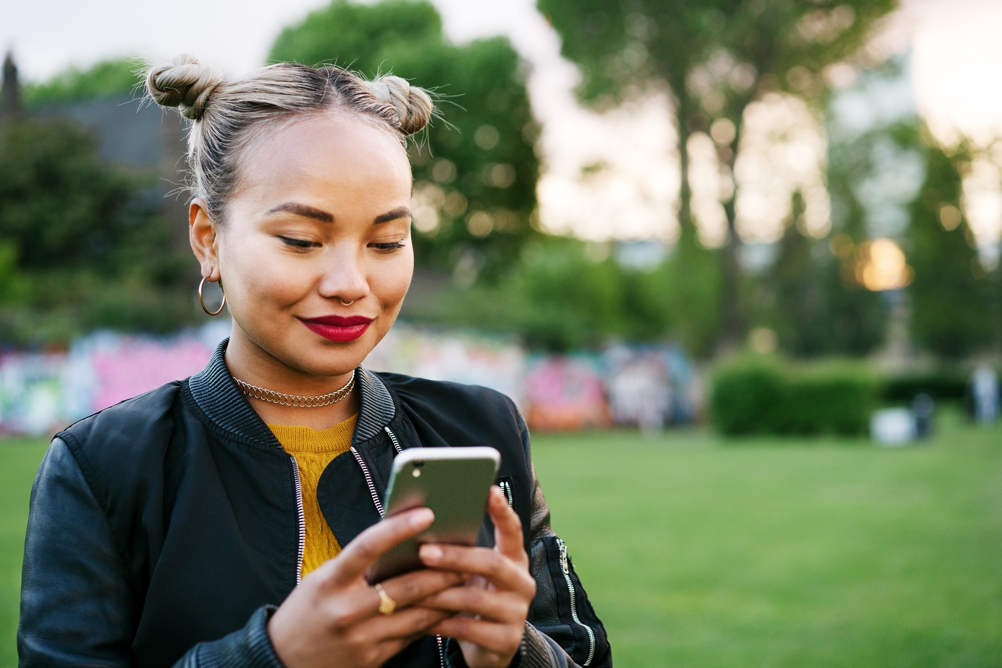 A woman with bunches in her hair in an outdoor setting smiles at her smartphone.