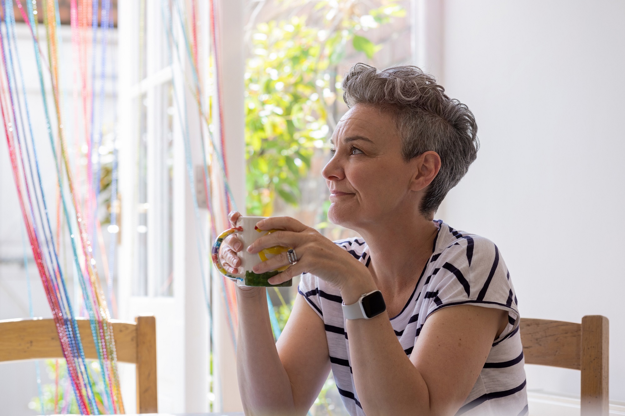 A woman with grey short hair holds a mug and looks out of a window in a brightly lit room.
