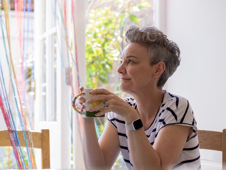A woman with grey short hair holds a mug and looks out of a window in a brightly lit room.