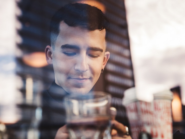 A young man with black hair and a hoop earring looks at his phone whilst sitting inside a glass fronted building.