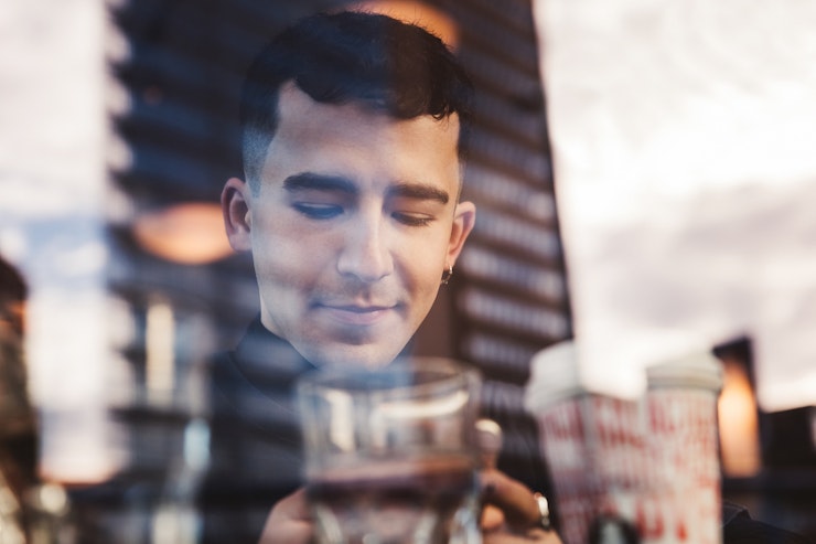 A young man with black hair and a hoop earring looks at his phone whilst sitting inside a glass fronted building.