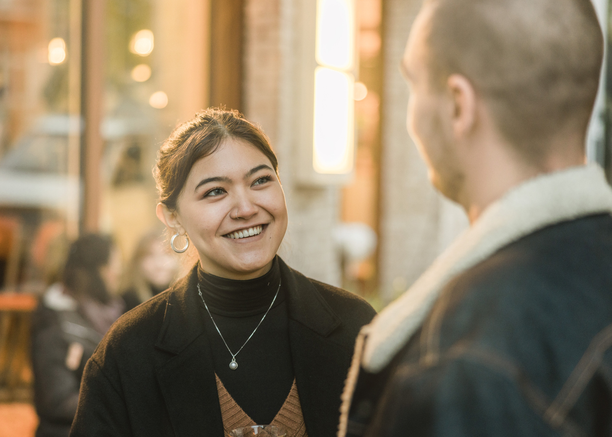 A young woman with hair pulled back smiles at a young man with close shaved hair in a cafe bar setting