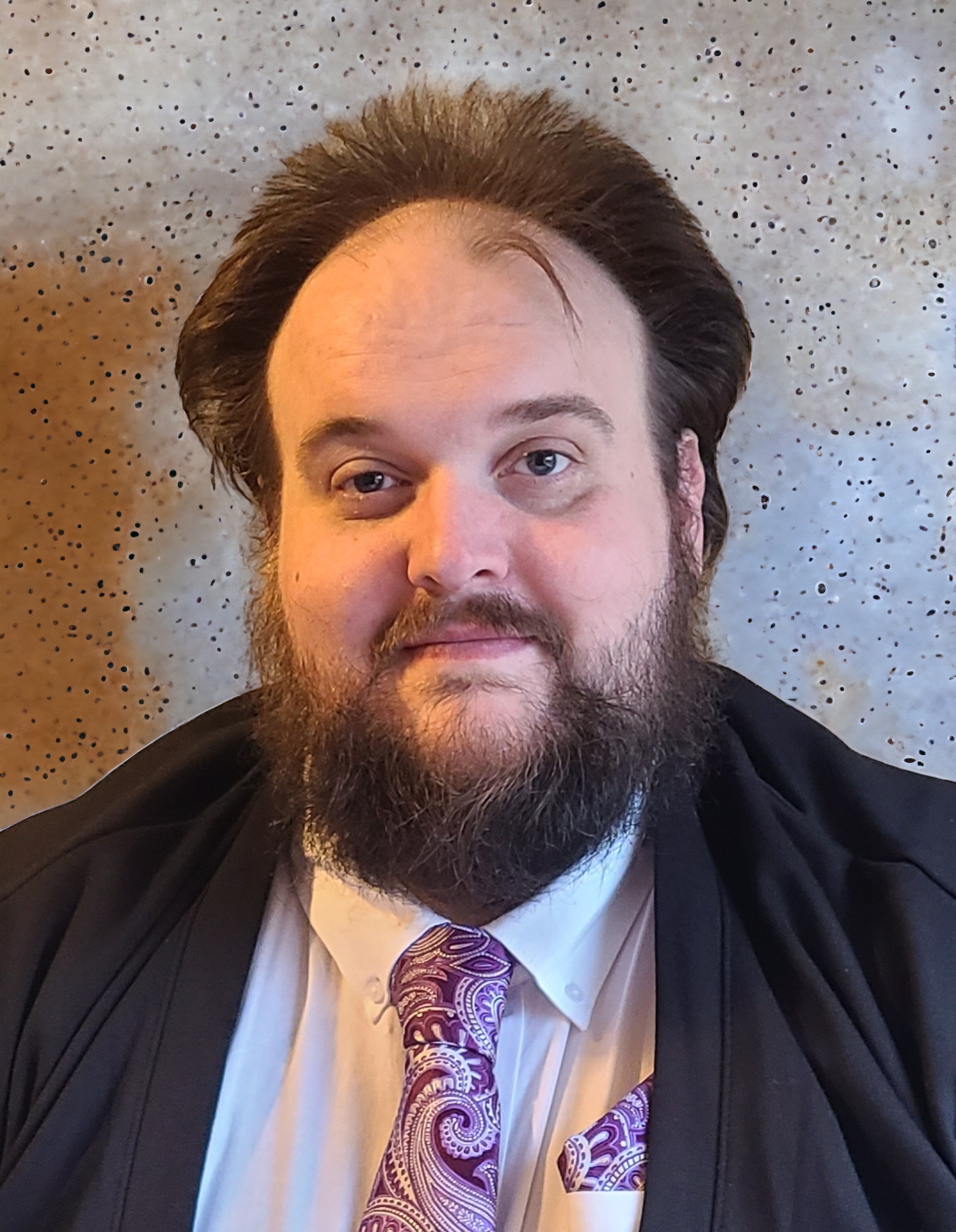 Alec smiles at the camera, wearing a suit and tie and standing indoors.