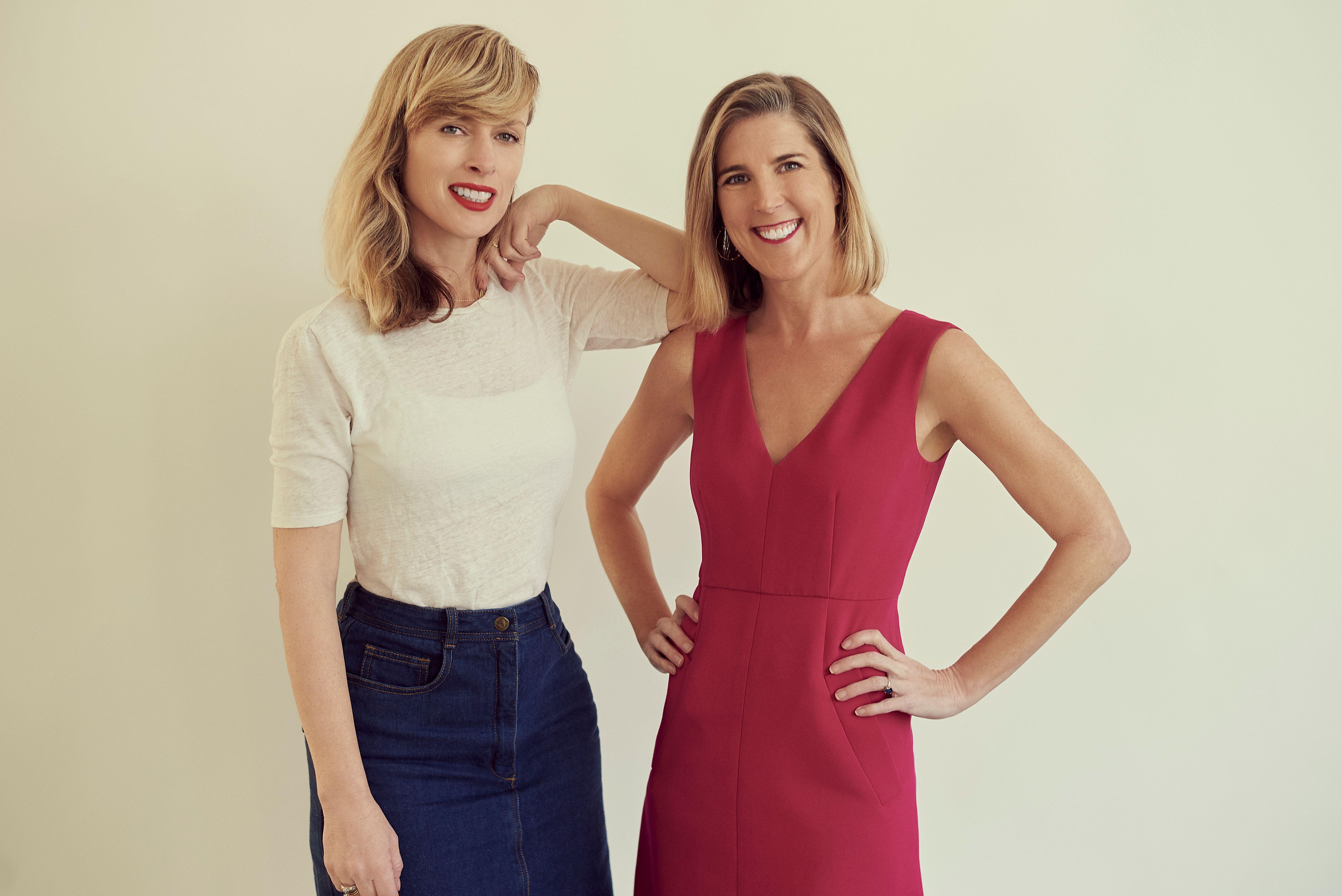 A woman in a red dress and a woman in a white top and black trousers lean on one another and smile.