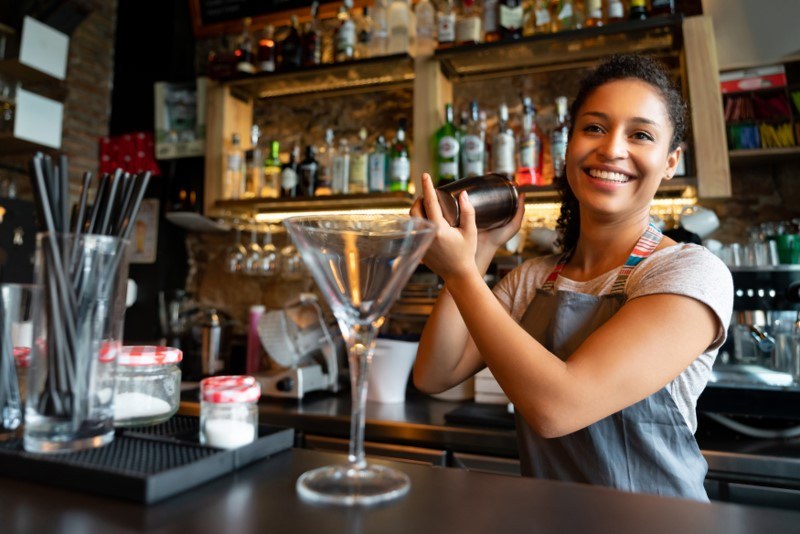 A bar woman wearing an apron smiles as she shames a drink.