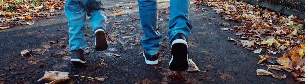 Two sets of legs walking in an autumnal street; one from a child and one from an adult.