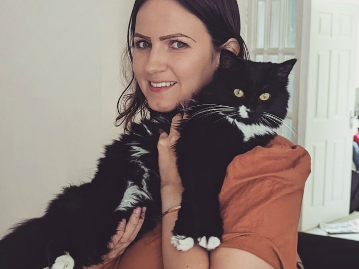 Cara stands smiling at the camera, holding a long-haired black and white cat.