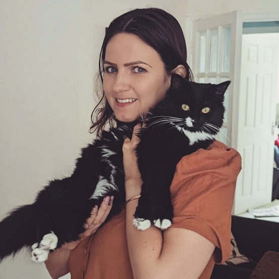 Cara stands smiling at the camera, holding a long-haired black and white cat.