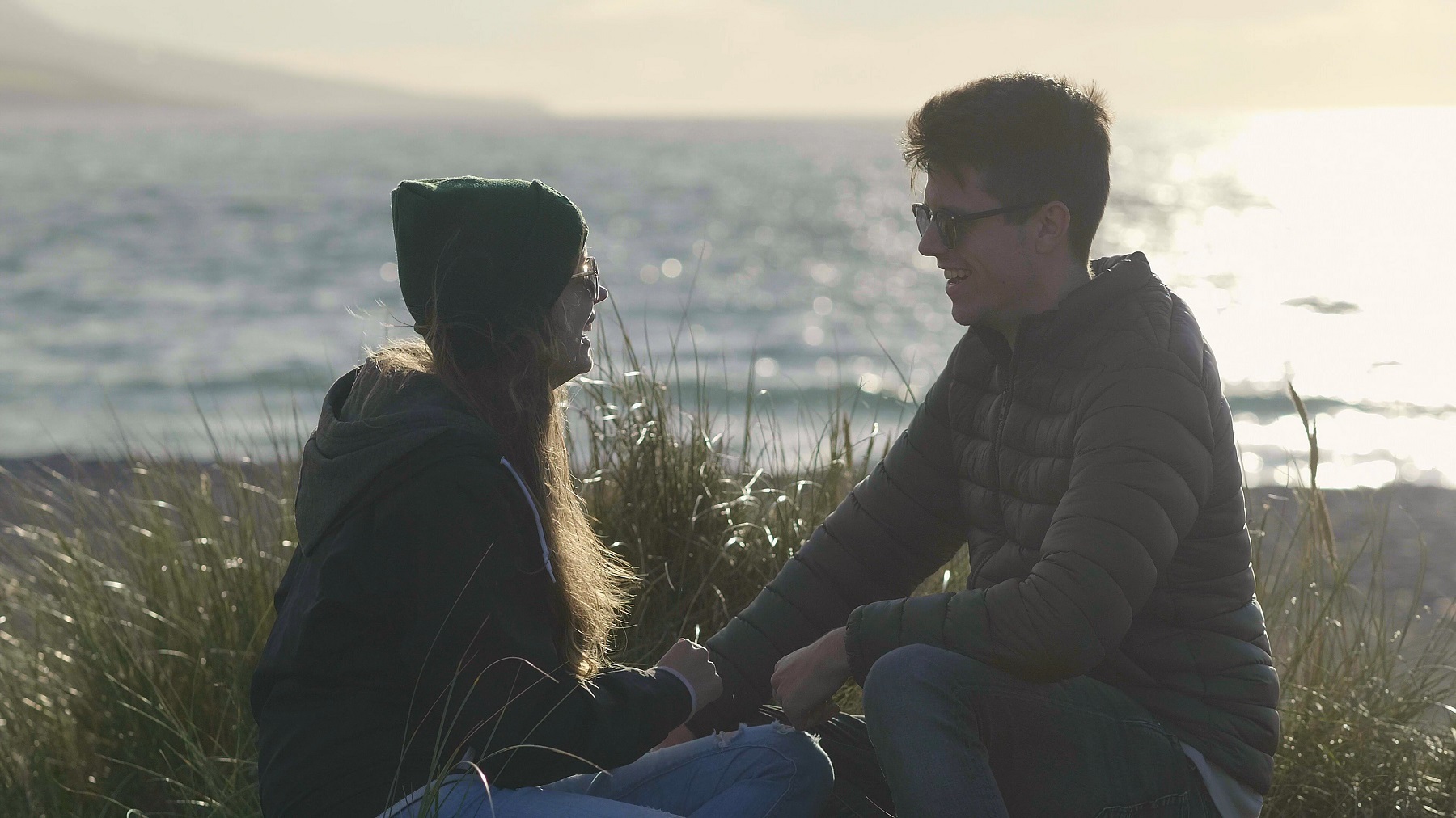 Couple laughing together as they sit cross-legged by a wintery beach at sunset.