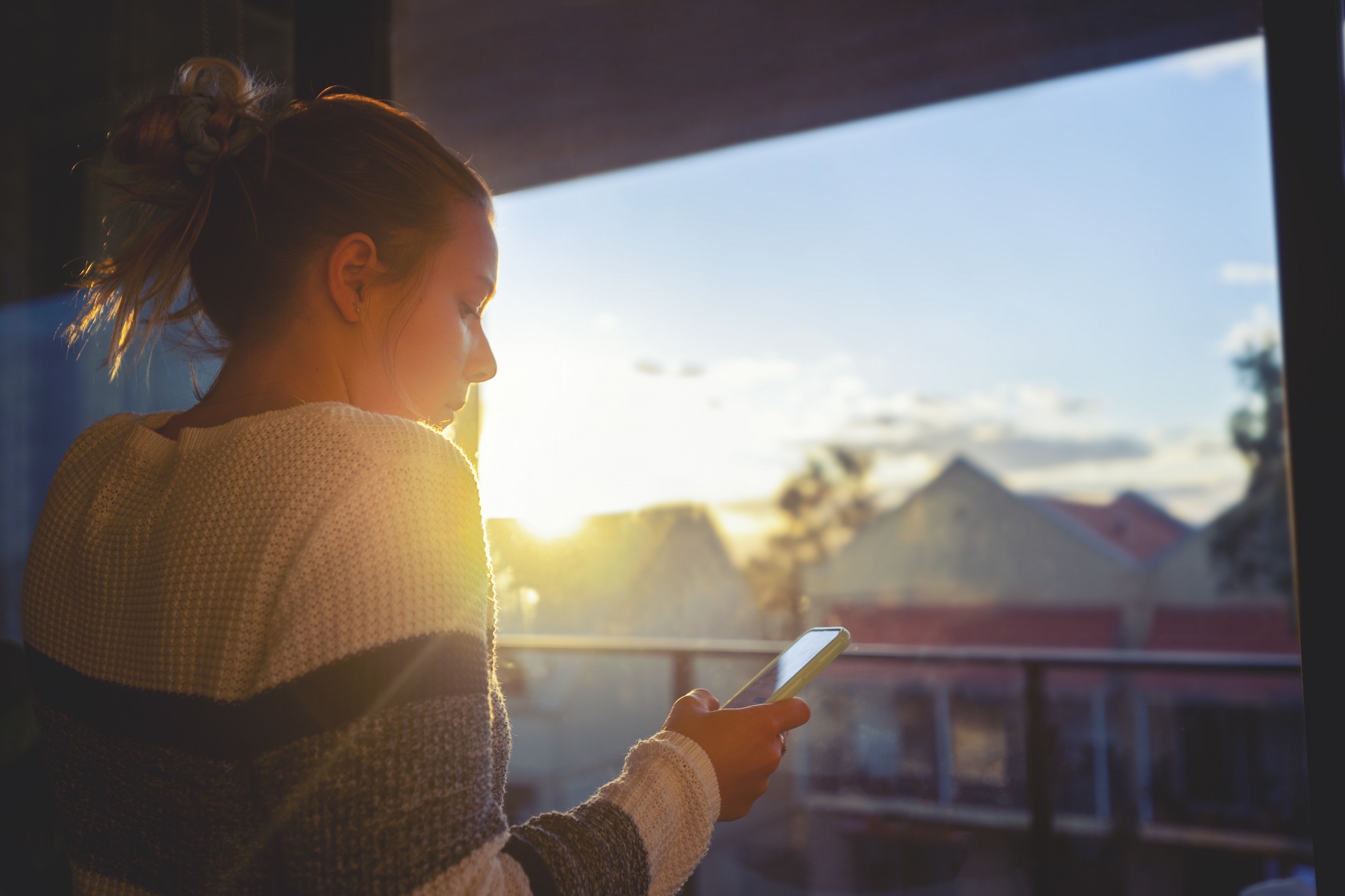 Girl looking through window at sunset using mobile phone