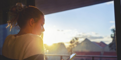 Girl looking through window at sunset using mobile phone