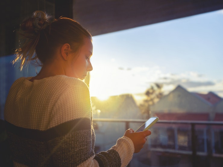 Girl looking through window at sunset using mobile phone