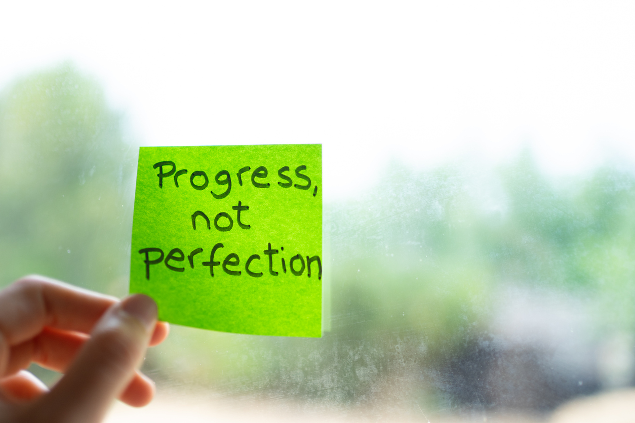 Hand is shown holding a green sticky note with progress not perfection written on it, in front of a window