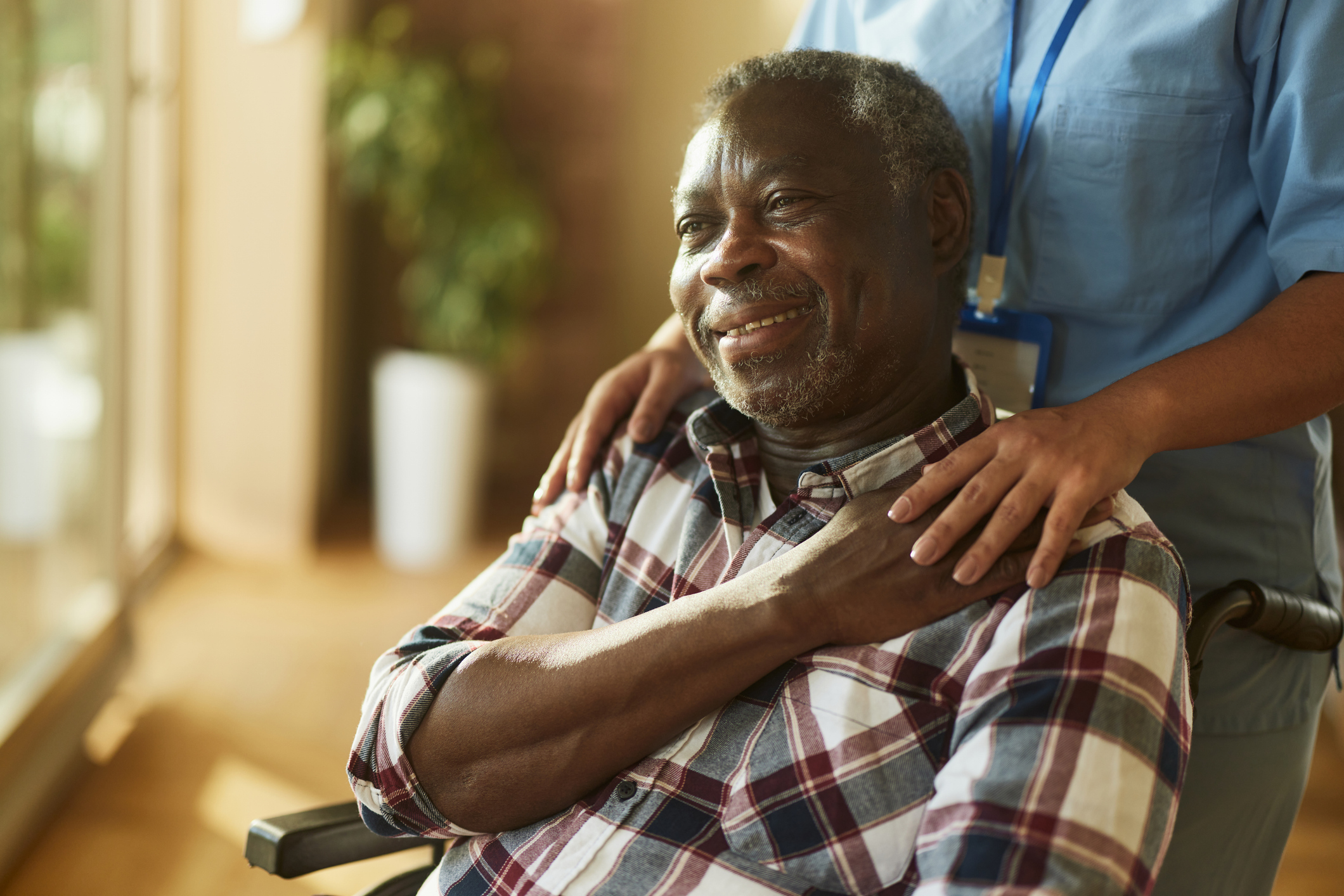 Happy African American mature man in a wheelchair at home looking around with support workers hands on shoulders