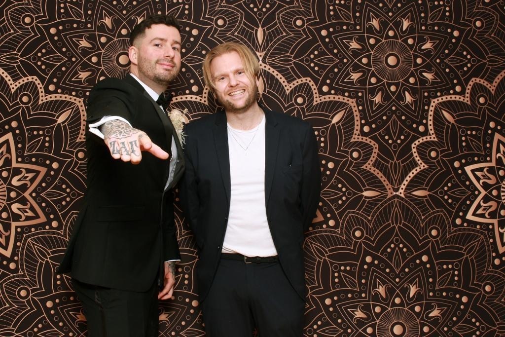 Two men wearing suits standing in front of an intricately patterned background smile and gesticulate.