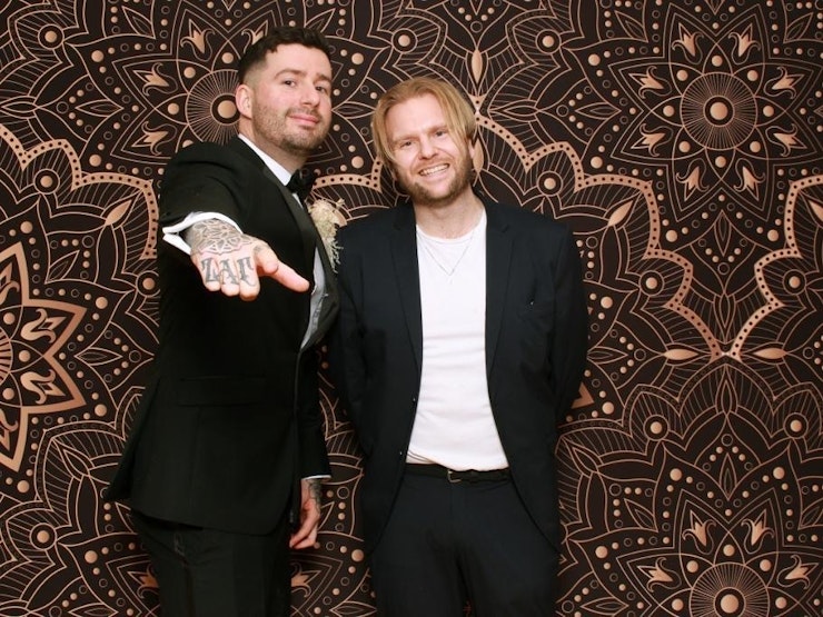 Two men wearing suits standing in front of an intricately patterned background smile and gesticulate.