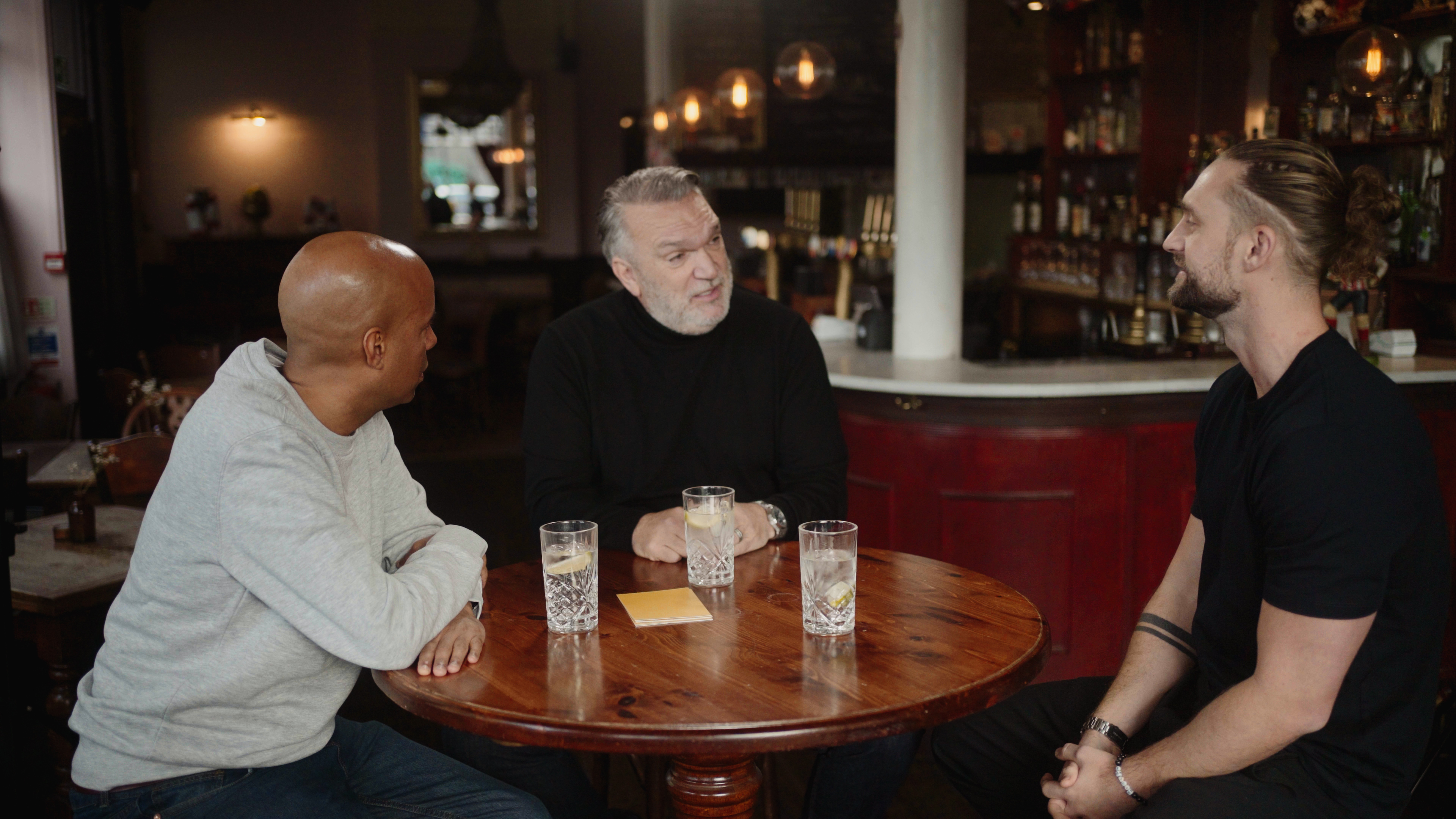 Three men sit in a dark pub with drinks.