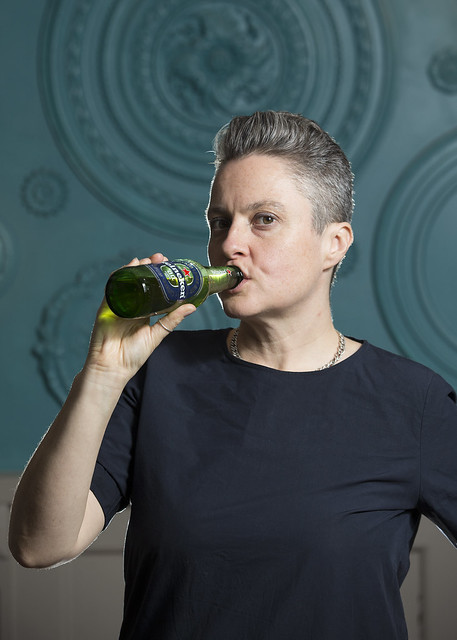 A woman in a black shirt with short, cropped grey hair swigs from a green glass bottle. Decorative background.