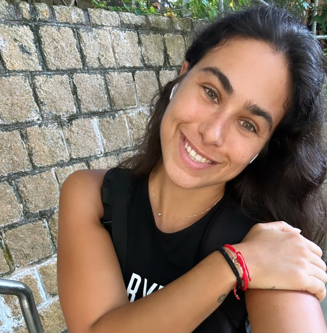 A woman with dark, straight hair hair smiles at the camera in front of a grey brick wall in an outdoor setting.