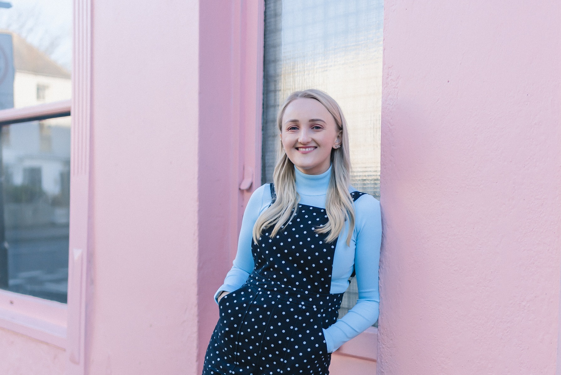 A woman wearing dungarees leans against a pink wall outdoors as she smiles at the camera.
