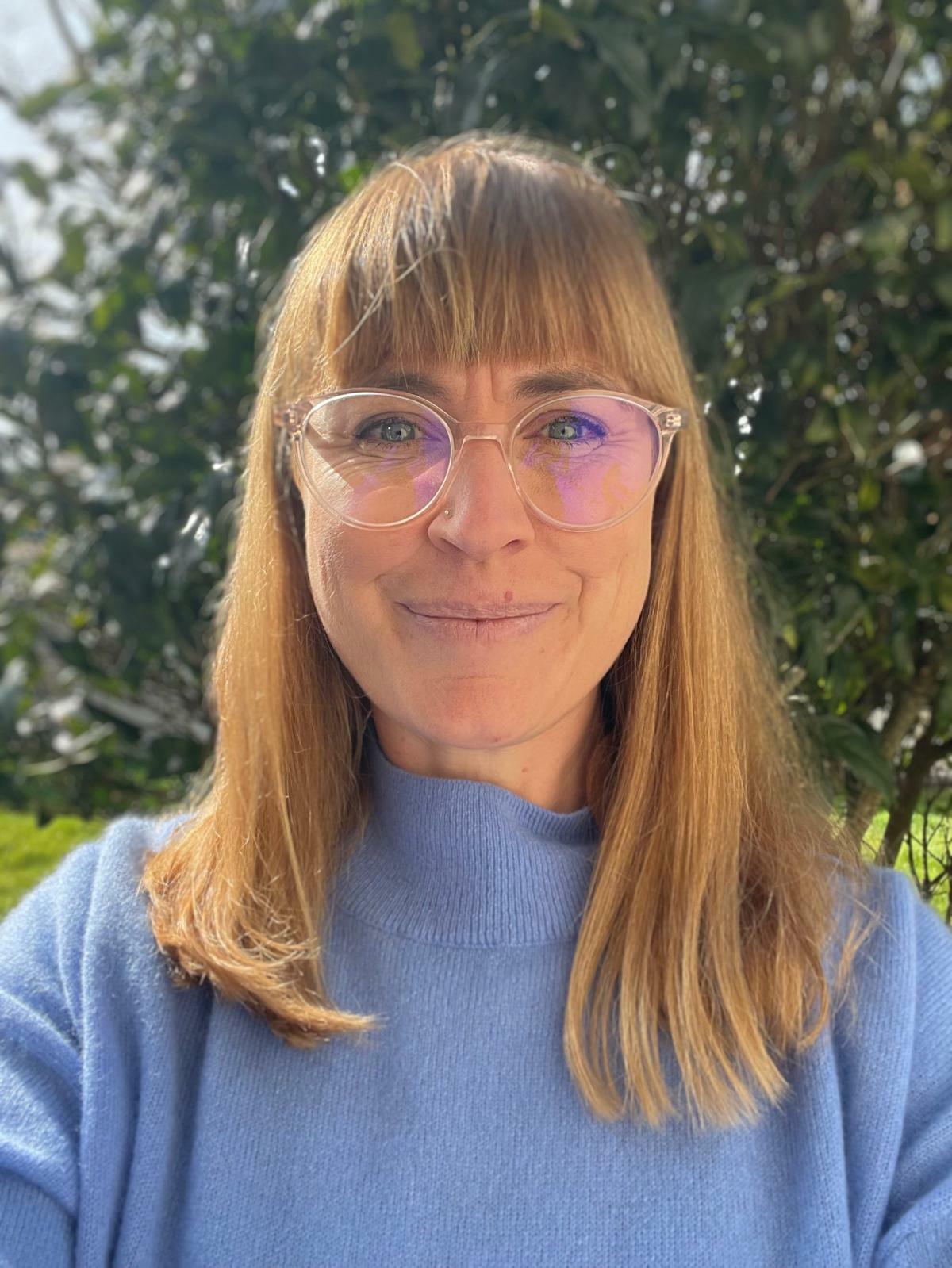 A woman wearing a blue jumper with round glasses on, smiles at the camera as she stands in a lush green outdoor space.