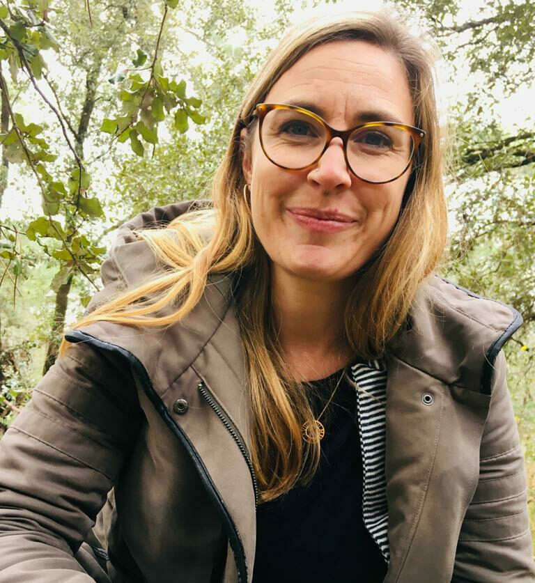 A blonde woman with glasses on in a forest smiling.