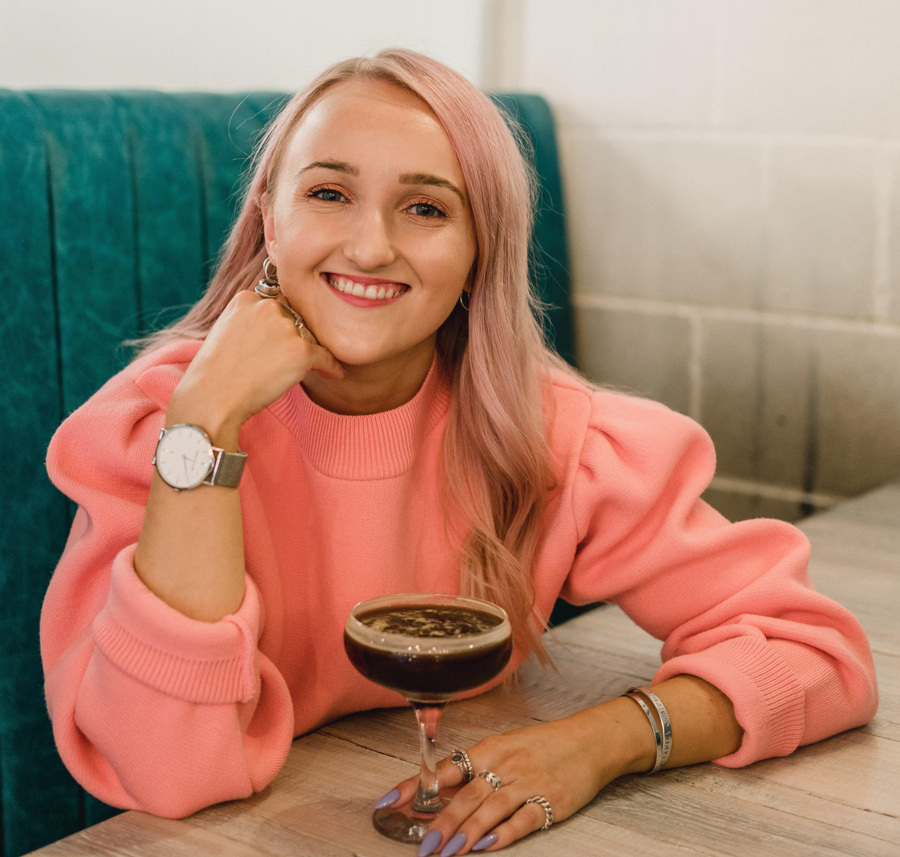 A woman with long, blond hair wearing a pink jumper sits at a table holding a drink and smiling.