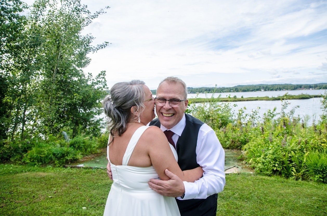 Older couple embrace and smile while wearing traditional wedding outfits in a lush, outdoors setting.