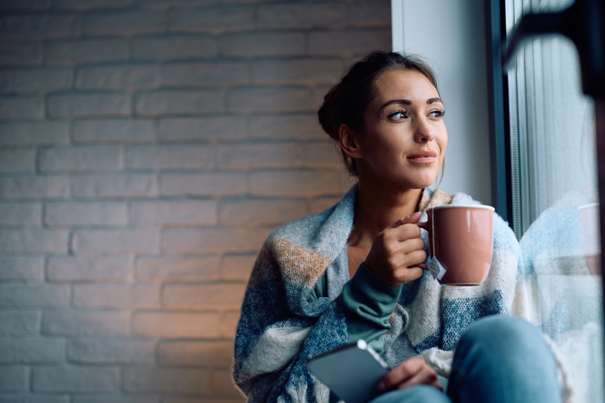 Image of a woman sitting on window sill drinking coffee at home while looking reflectively out of the window