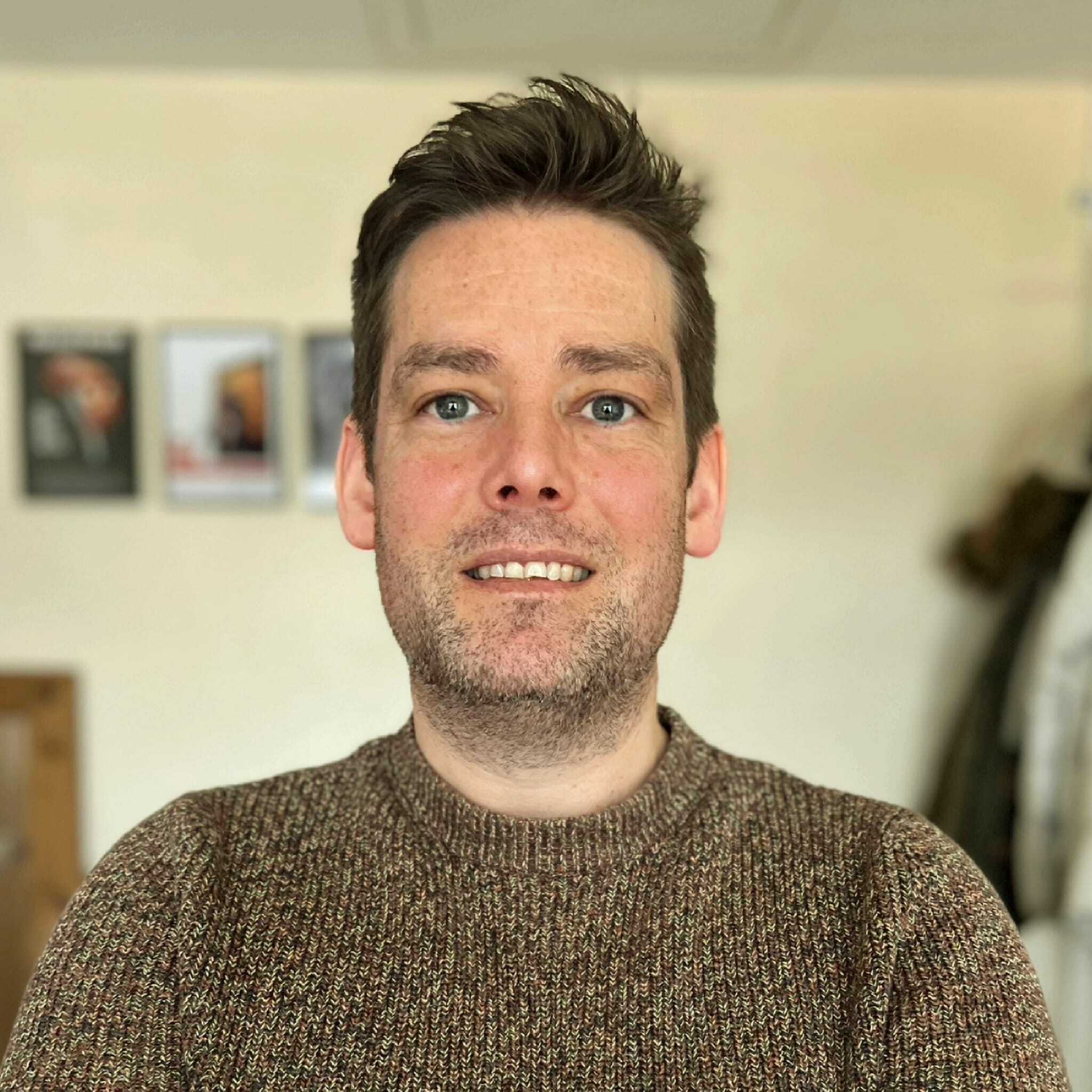 A man with cropped brown hair smiles at the camera in a homely environment.