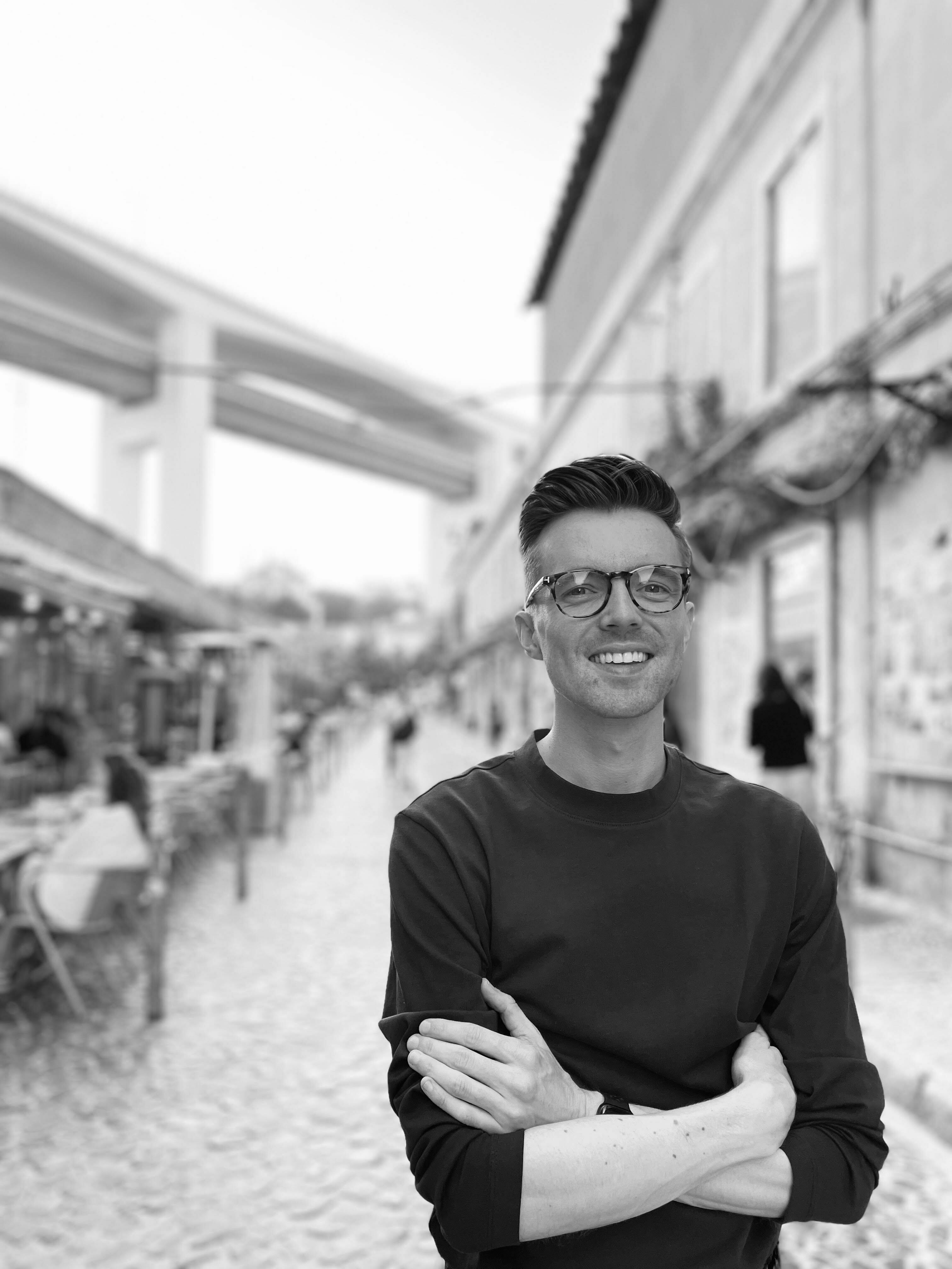 A man stands with his arms folded, grinning broadly, on a busy street where people sit outside at a restaurant.
