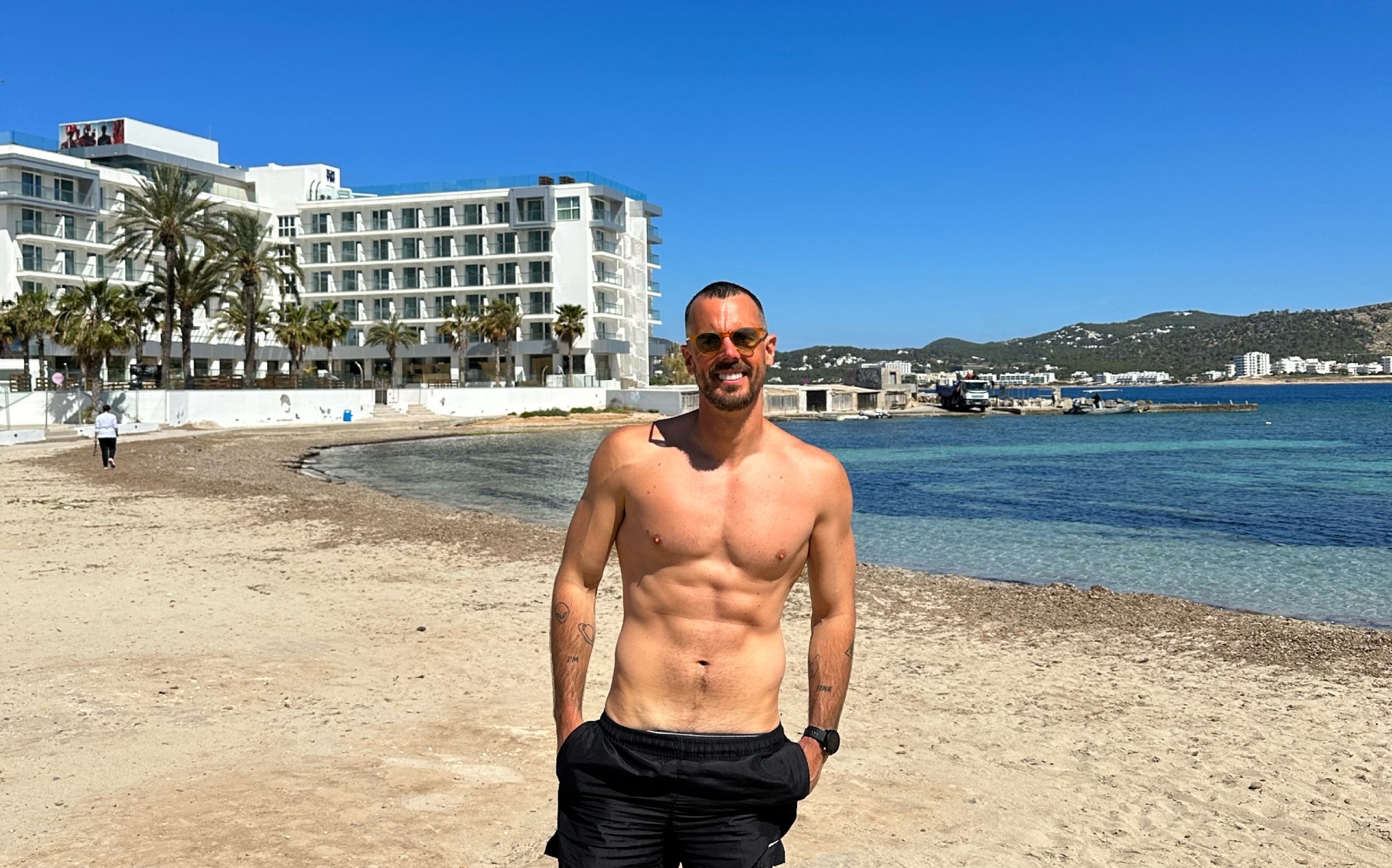 Man wearing swimming trunks and sunglasses on a beach smiles at the camera.