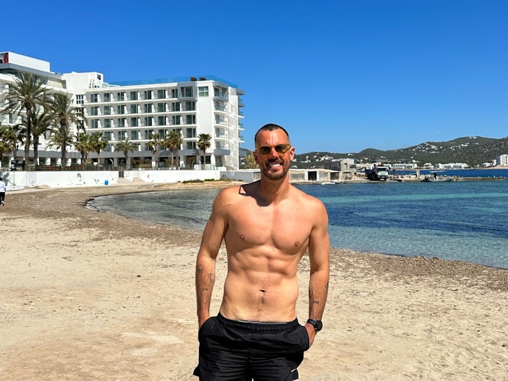 Man wearing swimming trunks and sunglasses on a beach smiles at the camera.