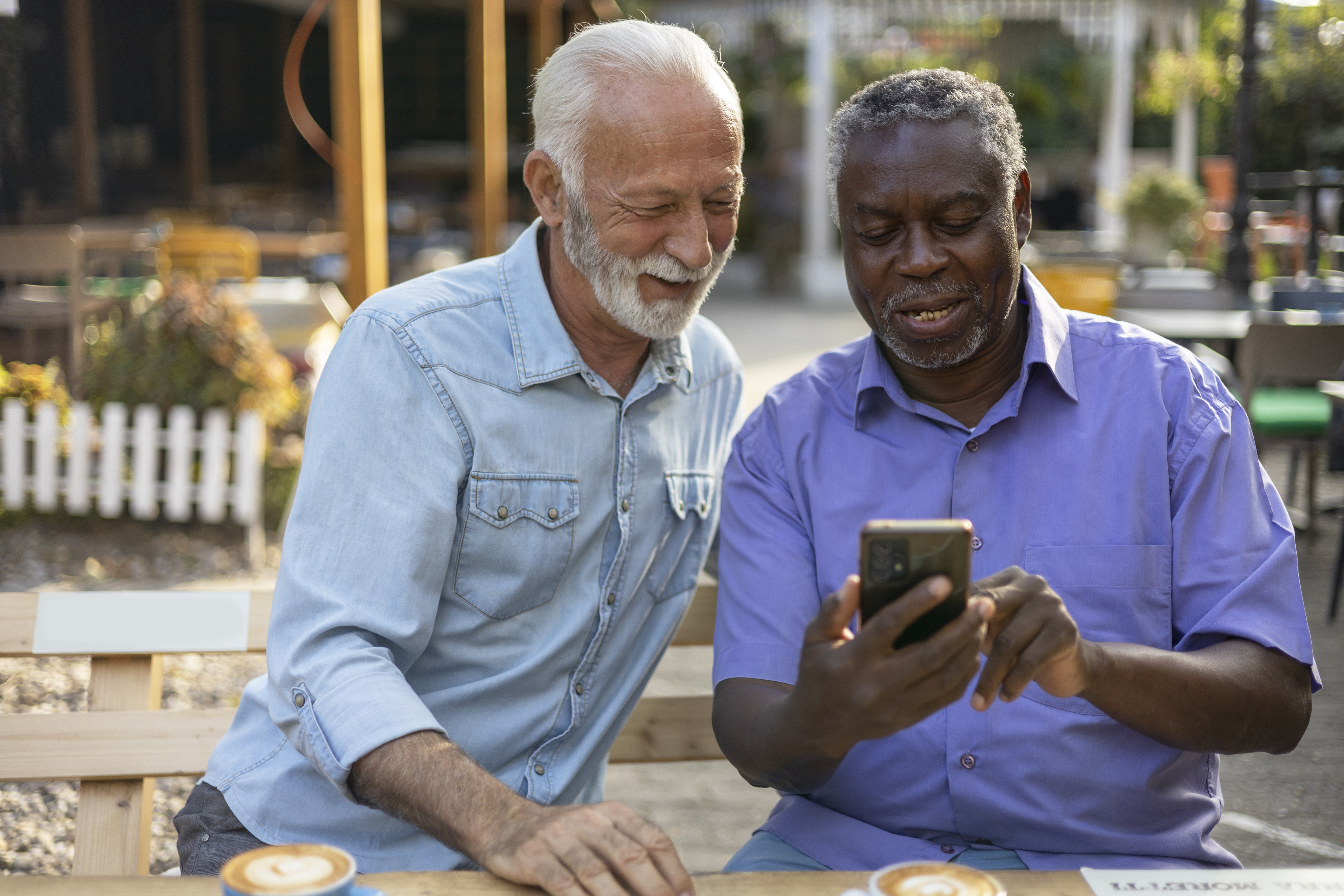 Two Diverse Senior Male Friends Using Smartphone While Sitting Outside at the Coffee Shop