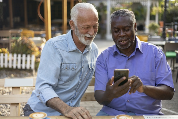 Two Diverse Senior Male Friends Using Smartphone While Sitting Outside at the Coffee Shop