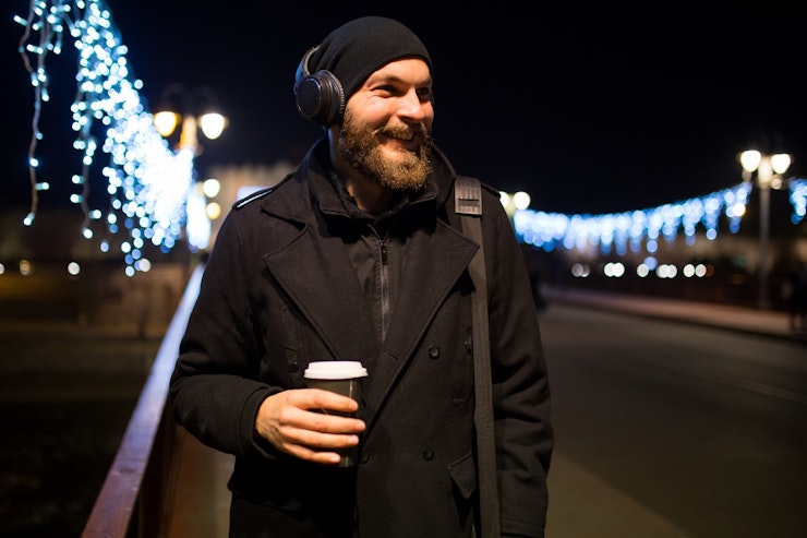 A smiling man in a hat on a cold night with festive lights behind him holds and coffee takeout.