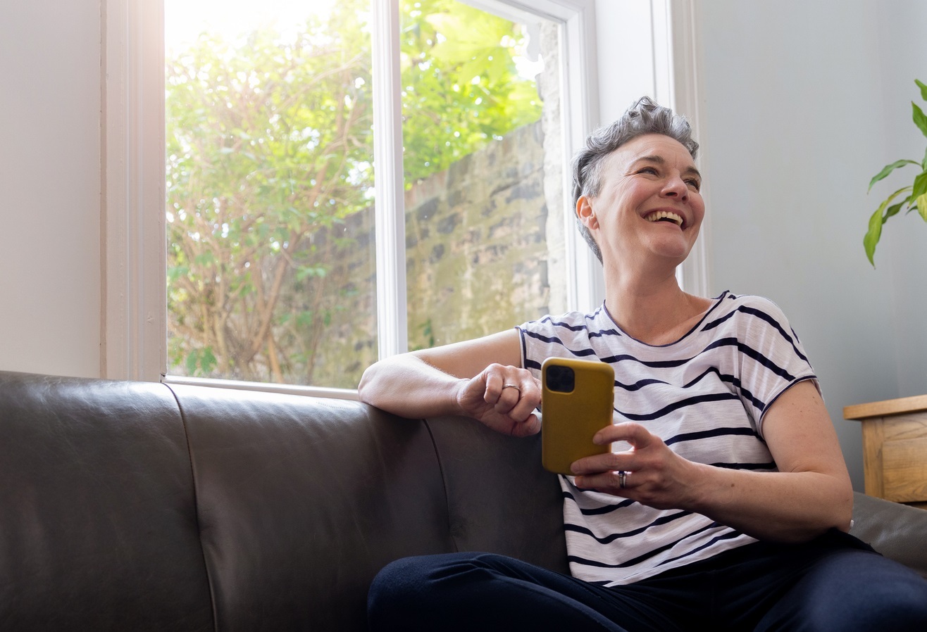 A smiling woman with grey, cropped hair sits on sofa using a smartphone in a yellow case.