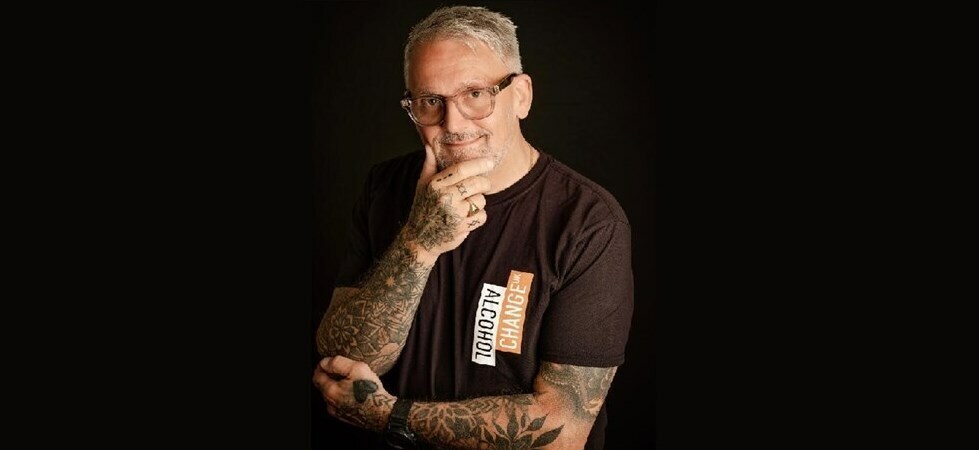 A man with grey hair and glasses clutches his chin whilst wearing and Alcohol Change UK T-shirt.