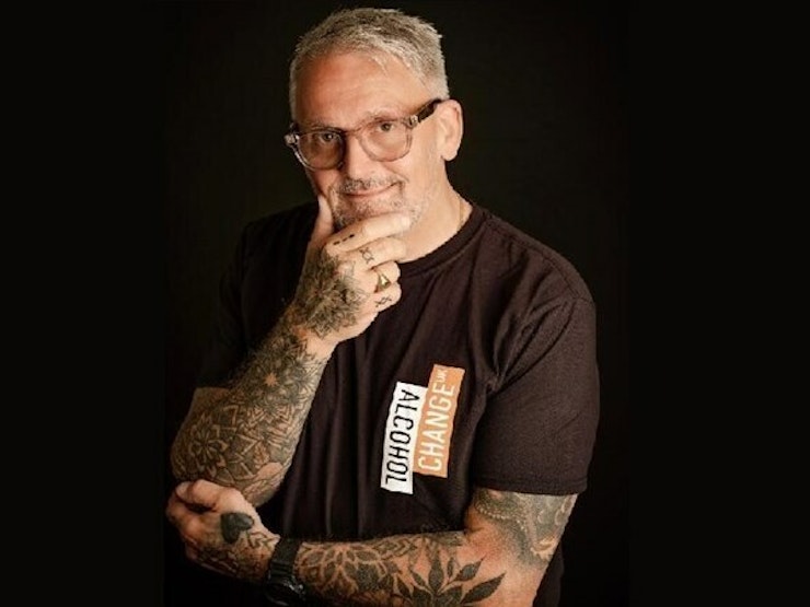 A man with grey hair and glasses clutches his chin whilst wearing and Alcohol Change UK T-shirt.