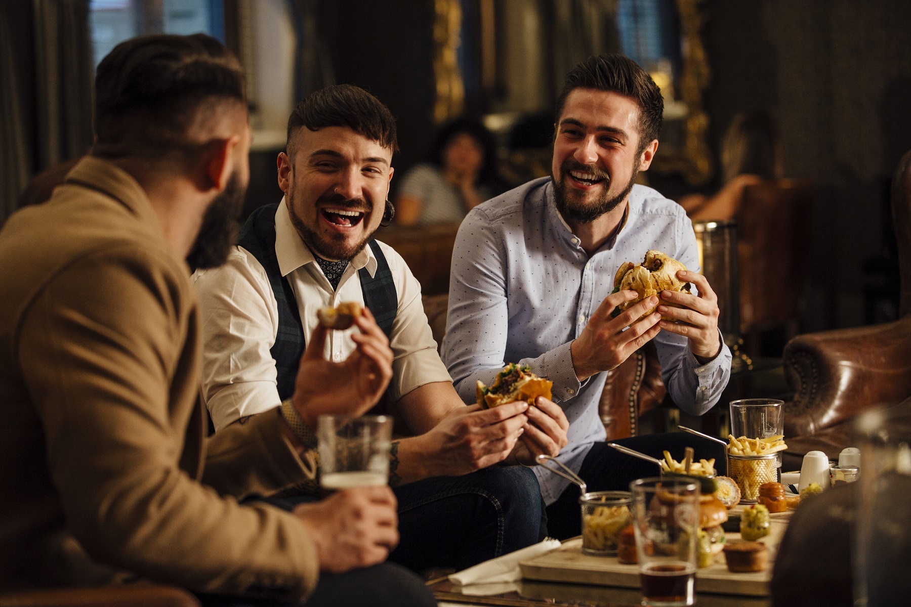Three men in a pub eat burgers and have drinks whilst they laugh and joke together.