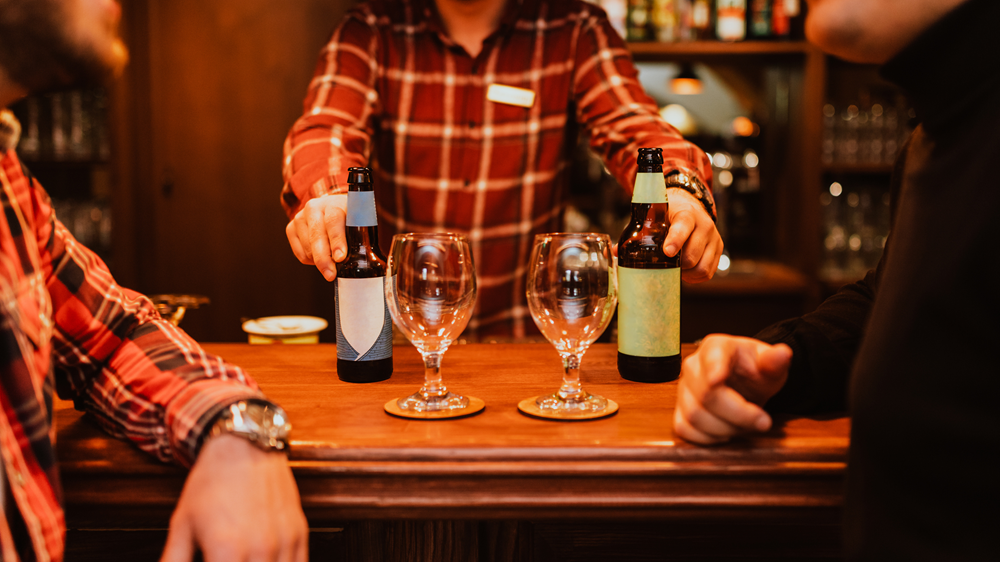 Two men at a bar with empty glasses about to drink from two beer bottles provided by a barman.
