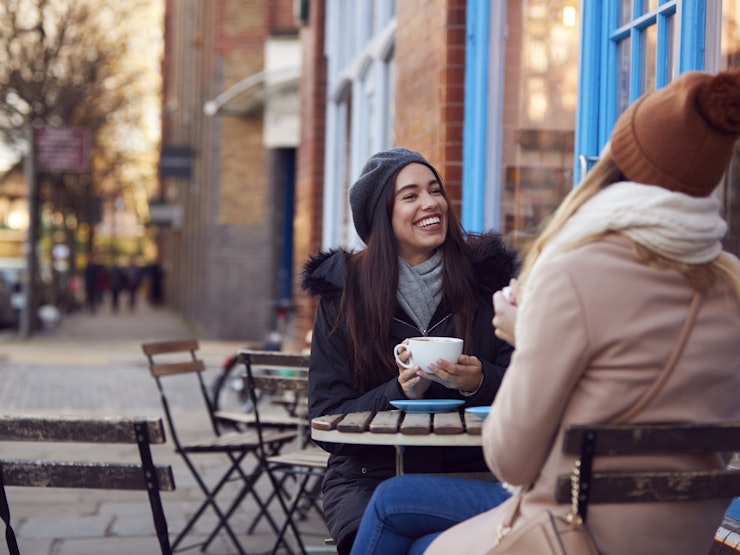 Two women wearing wooly hats sit outside with coffee cups smiling and laughing.
